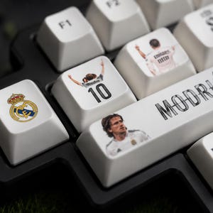 May include: A black mechanical keyboard with custom white keycaps featuring Real Madrid graphics. Keycaps display the team logo, the number 10, a portrait of Luka Modrić, the text MODRIĆ, and the Emirates Fly Better logo in black and white.