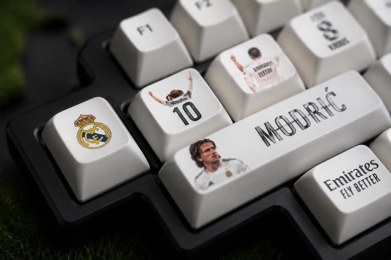May include: A black mechanical keyboard with custom white keycaps featuring Real Madrid graphics. Keycaps display the team logo, the number 10, a portrait of Luka Modrić, the text MODRIĆ, and the Emirates Fly Better logo in black and white.