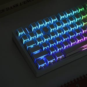 May include: A white mechanical keyboard with illuminated keys. The keys are lit with a gradient of colors, transitioning from blue to purple and green. The keyboard has a compact layout with function keys and a number row.