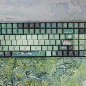 May include: A mechanical keyboard featuring a color scheme of light and dark green keys. Several keys display detailed illustrations of forest spirits and natural landscapes. The spacebar and other select keys feature printed scenes with characters.