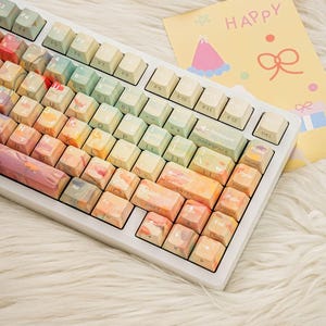 May include: A white mechanical keyboard with colorful keycaps. The keycaps have a watercolor design in shades of orange, pink, green, and blue. A yellow card with the word "HAPPY" and a party hat illustration is in the background.