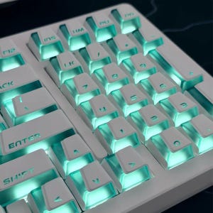 May include: A white computer keyboard featuring a numeric keypad and function keys with translucent sides. The keys are illuminated from beneath with a bright teal glow, highlighting the white keycaps and the dark teal lettering on each key.