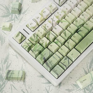 May include: A white mechanical keyboard with keycaps featuring a green and white leaf pattern. The keyboard has standard keys, including function keys, letters, numbers, and special keys like Shift, Ctrl, and Alt.