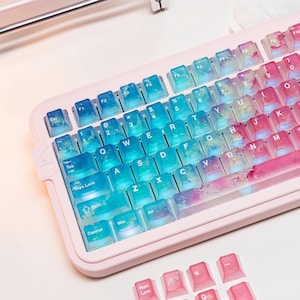 May include: A pink mechanical keyboard with translucent keycaps. The keycaps transition from blue to pink, with a watercolor effect. Additional pink keycaps are at the bottom. The keyboard has standard QWERTY layout with function keys.