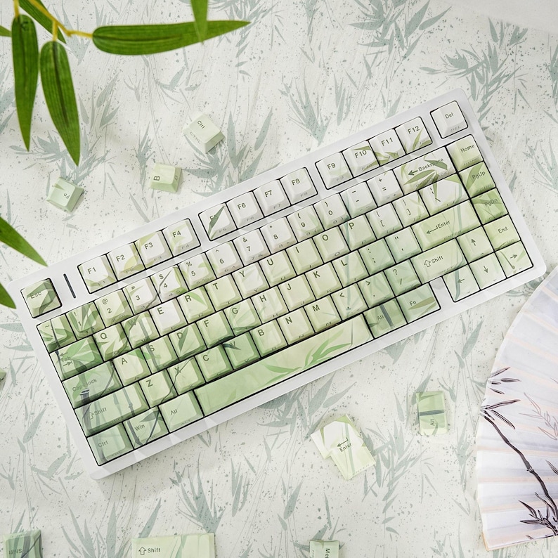 May include: A white mechanical keyboard with keycaps featuring a green bamboo leaf design. The keyboard has a standard layout with function keys and a number pad. The background has a bamboo leaf pattern.
