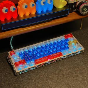 May include: A backlit mechanical keyboard with blue and red keycaps. The keyboard has a silver frame with a geometric design. The keycaps are transparent, allowing the blue backlighting to shine through. The keyboard also has a red space bar with the text "E.F.S.F."
