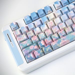 May include: A mechanical keyboard featuring a white frame and custom keycaps decorated with a soft watercolor floral pattern in shades of pink, blue, and green. The top row of keys transitions into a clear blue sky design with white clouds.