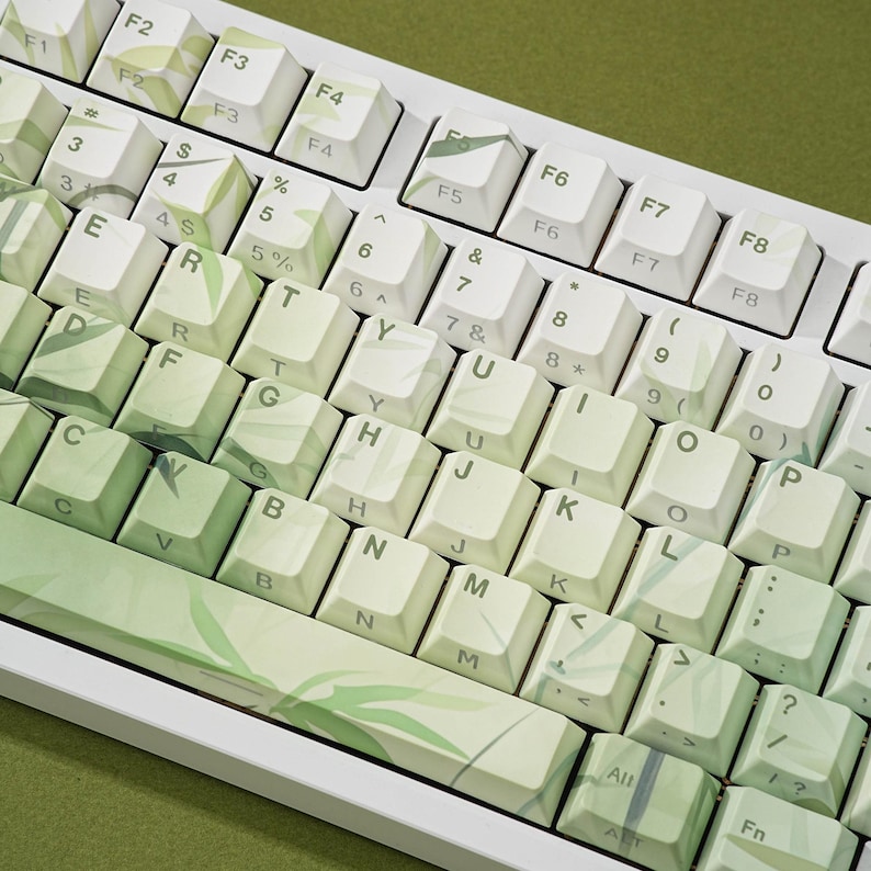 May include: A close-up of a white mechanical keyboard with green and white keycaps. The keycaps have a leaf pattern design. The keyboard is on a green surface.