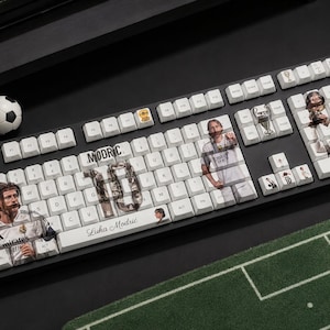 May include: A black mechanical keyboard with white keycaps featuring custom graphics of Luka Modrić, the number 10, and soccer-themed icons. The text "MODRIC" and "Luka Modrić" is printed on the keys, alongside images of the player in a white jersey.