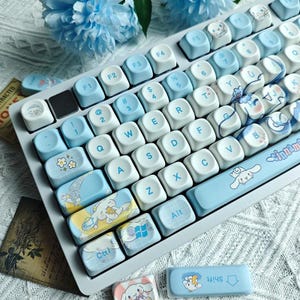 May include: A light blue and white keyboard with custom keycaps. The keycaps feature various designs, including floral patterns and cartoon characters. The keyboard has a light blue space bar with the word "cinnamon" and a cartoon character.