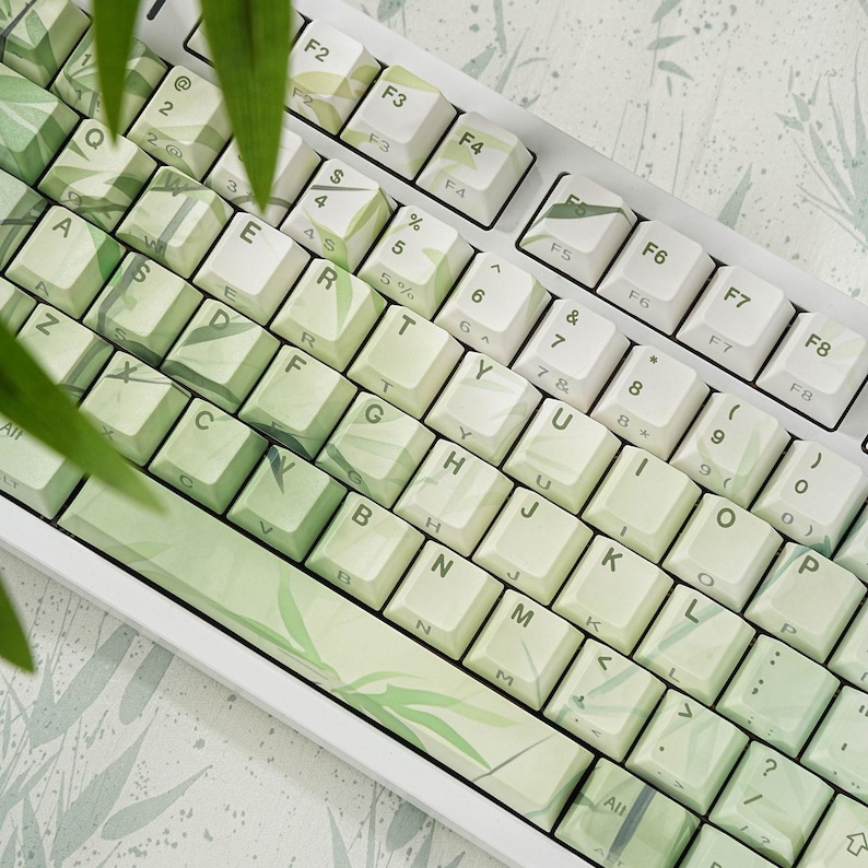 May include: A white mechanical keyboard with keycaps featuring a green and white bamboo leaf design. The keyboard has a standard QWERTY layout with function keys and number pad. The keycaps have a gradient effect, transitioning from light to dark green.
