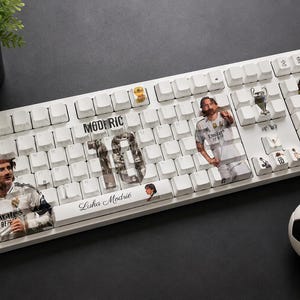 May include: A white mechanical keyboard featuring custom keycaps with images of Luka Modrić, the number 10, and the text "MODRIĆ" and "Luka Modrić". The keyboard includes small decorative icons of a trophy and a soccer ball design on specific keys.