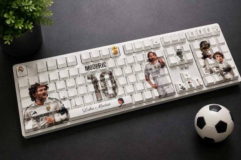 May include: A white mechanical keyboard featuring custom keycaps with images of Luka Modrić, the number 10, and the text "MODRIĆ" and "Luka Modrić". The keyboard includes small decorative icons of a trophy and a soccer ball design on specific keys.