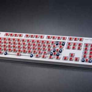 May include: A white mechanical keyboard base without keycaps, featuring numerous red and blue switches exposed across the layout. A circular volume knob is positioned on the top right corner, and a subtle blue light glows from the side edge.