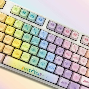 May include: A pastel-colored keyboard with bear-shaped keycaps. The keys are arranged in a standard layout, with function keys and a space bar that reads "SWEET BEAR." The keycaps feature various symbols and characters.