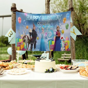 May include: A birthday party scene with a banner that reads "Happy Birthday Olivia 5". The banner features characters from the movie Frozen, balloons, and gift boxes. A cake with a car on top and other food items are on a table.