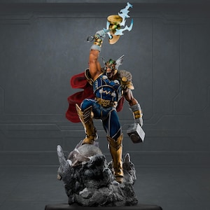 Könnte beinhalten: Detaillierte Statue eines Superhelden in dynamischer Pose, der eine leuchtende Axt und einen Hammer hält. Die Figur trägt ein blau-goldenes Kostüm mit rotem Umhang und steht auf einem felsigen Sockel. Die Statue hat einen wilden Gesichtsausdruck.