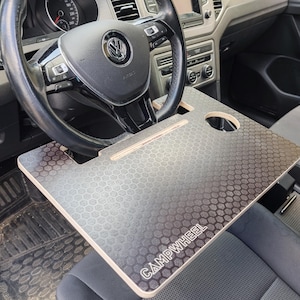 Könnte beinhalten: Ein hölzerner Schreibtisch für das Lenkrad eines Autos mit einem Wabenmuster und dem Text "CAMPWHEEL". Der Schreibtisch wird auf dem Lenkrad platziert und ist als Tisch konzipiert. Der Innenraum des Autos umfasst ein schwarzes Lenkrad und Armaturenbrett.