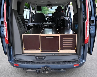 May include: A dark blue van with the rear doors open, revealing a custom-built wooden storage system. The system includes drawers and compartments, likely for equipment or tools. The van's interior is visible, with seats and a dashboard.