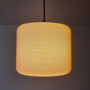 Op de afbeelding: Een cilindervormige hanglamp met een geweven lampenkap. De lampenkap is verlicht en geeft een warme, gele gloed. De lamp hangt aan een zwart snoer tegen een neutrale achtergrond.