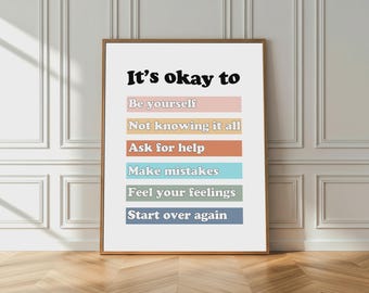May include: A framed print with the text "It's okay to" at the top, followed by phrases like "Be yourself" and "Start over again" on colored blocks. The print has a wooden frame and is displayed against a white wall with a wooden floor.