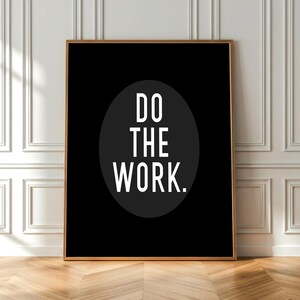 May include: A framed black and white motivational art print. The artwork features the phrase "DO THE WORK" in bold white letters within a black oval against a black background. The frame is gold-toned, and the print is displayed against a white wall.
