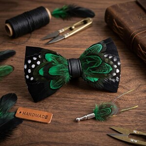 May include: A handmade black feather bow tie with green accents and white polka dots, featuring a black leather band. A matching feather lapel pin is also present. The bow tie and pin are displayed with sewing supplies on a wooden surface.