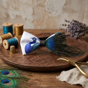 Puede incluir: Una pajarita blanca con un diseño de pavo real, con plumas azules y verdes. La pajarita está sobre una bandeja de madera con utensilios de costura y plumas de pavo real. La pajarita tiene una banda negra.