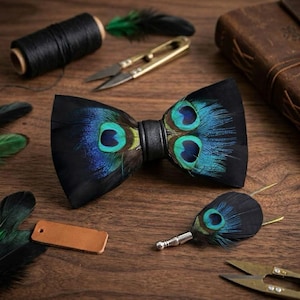Puede incluir: Una pajarita y un prendedor negros con detalles de plumas de pavo real. La pajarita presenta una banda de cuero negro y detalles de plumas azules y verdes vibrantes. También se ven unas tijeras doradas y una bobina de hilo negro.