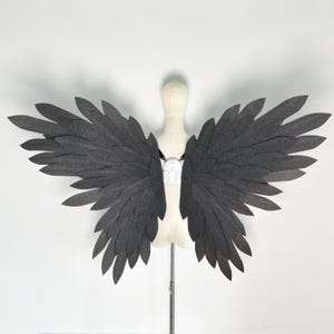 Può includere: Ali in feltro grigio scuro con un design a piume a strati, esposte su un manichino. Le ali sono attaccate con cinghie nere e un fermaglio bianco. Un accessorio di costume.