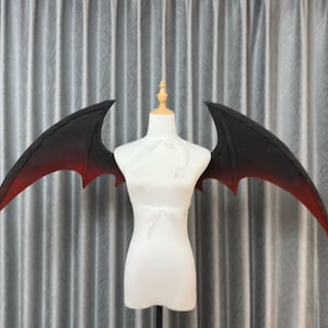 May include: A pair of black and red ombre cosplay wings, displayed on a white dress form. The wings have a pointed shape and a gradient effect, transitioning from black to red. The dress form is set against a backdrop of gray curtains.