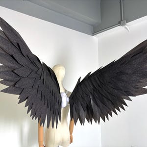 Könnte beinhalten: Große, dunkelgraue Kostümflügel mit einem geschichteten Federdesign. Die Flügel werden auf einer Schaufensterpuppe vor einer weißen Wand ausgestellt. Die Flügel bestehen aus einem strukturierten Material und sind weit ausgebreitet.