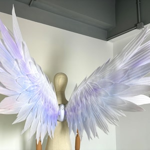 May include: Large, white angel wings with a soft, iridescent lavender and blue gradient. The wings are attached to a mannequin and have a layered, feather-like design. The wings are a costume or decorative item.