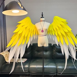 May include: A pair of large, yellow and white angel wings displayed on a mannequin. The wings are made of layered, feather-like material. The mannequin is on a black stand, and the background includes a green sofa and a decorative avocado pillow.