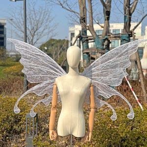 May include: A pair of large, translucent fairy wings with a glittery, iridescent finish. The wings are attached to a mannequin, showcasing their delicate, detailed design. The wings are a light, airy accessory.