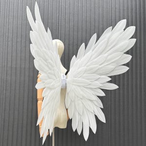 May include: White angel wings with layered feather design. The wings are attached to a white harness. The wings are displayed on a mannequin against a gray background. The wings are a costume accessory.
