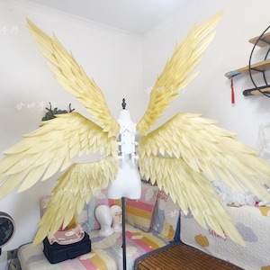 May include: Large, pale yellow angel wings displayed on a white mannequin. The wings are made of layered, feather-like material, with a central spine and multiple tiers of individual feathers. The wingspan is wide, and the overall effect is ethereal.
