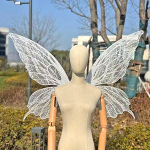 Peut inclure: Une paire d'ailes de fée blanches scintillantes avec des détails de veines complexes et des accents de paillettes. Les ailes sont présentées sur un mannequin, mettant en valeur leur design délicat et leur qualité éthérée. Parfait pour les soirées costumées ou les événements à thème.