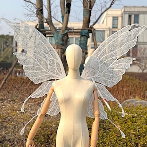 Może przedstawiać: Para delikatnych, białych skrzydeł wróżki ze skomplikowanymi koronkowymi detalami, prezentowanych na manekinie. Skrzydła mają kształt motyla i są przymocowane do pleców manekina. Tło przedstawia drzewa i budynek.
