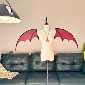May include: A mannequin with a necklace and a pink pendant stands in front of a wall with red bat wings. A green leather couch with a floral blanket is in front of the mannequin. A large, black lamp hangs from the ceiling.