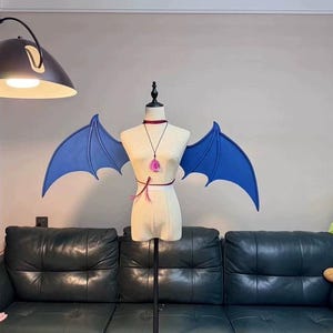 May include: A mannequin displays a pair of blue bat wings, a choker, and a pendant. The wings are a vibrant blue and are attached to the mannequin's back. The mannequin is on a stand in front of a dark green couch.