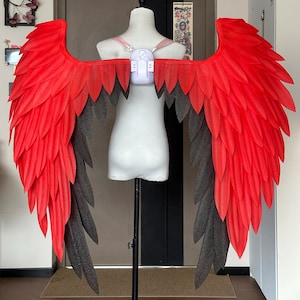 May include: Large, vibrant red and black costume wings. The wings are attached to a white harness, designed to be worn on the back. The feathers are layered, creating a textured appearance. The wings are displayed on a mannequin.
