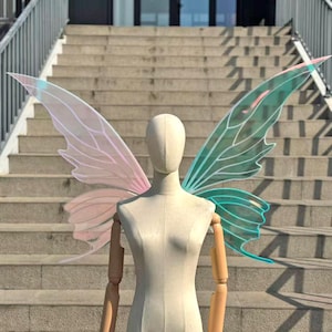 Puede incluir: Un par de alas de hada iridiscentes, exhibidas en un maniquí. Las alas presentan un degradado de rosa y turquesa, con delicados detalles en forma de venas. El maniquí está colocado frente a una escalera exterior.