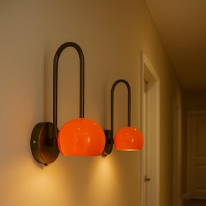 May include: Two modern wall sconces with orange globe shades and dark metal arched arms. The sconces are mounted on a light-colored wall, casting a warm glow. The design is minimalist and stylish, suitable for interior lighting.