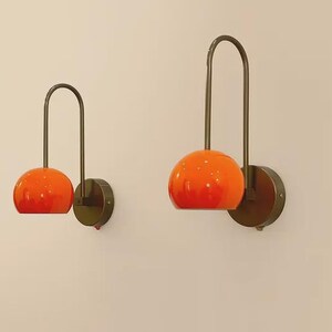 May include: Two wall sconces with orange globe shades and dark metal arched arms. The sconces are mounted on round, dark metal bases. The shades have a gradient effect, transitioning from a lighter orange at the top to a deeper orange at the bottom.