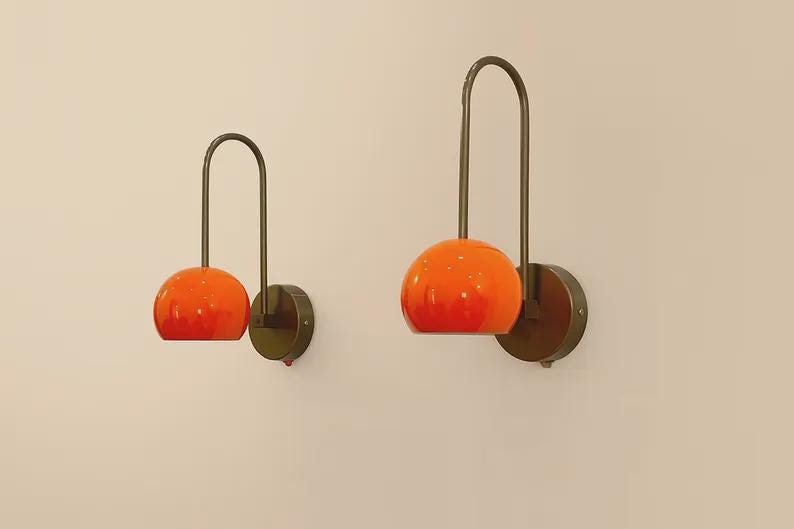 May include: Two wall sconces with orange globe shades and dark metal arched arms. The sconces are mounted on round, dark metal bases. The shades have a gradient effect, transitioning from a lighter orange at the top to a deeper orange at the bottom.