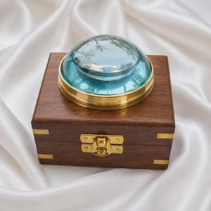 May include: A square dark brown wooden box featuring brass corner accents and a central brass latch. Mounted on the lid is a circular, clear glass dome with a blue-tinted base and a polished brass rim, creating a reflective, layered appearance.