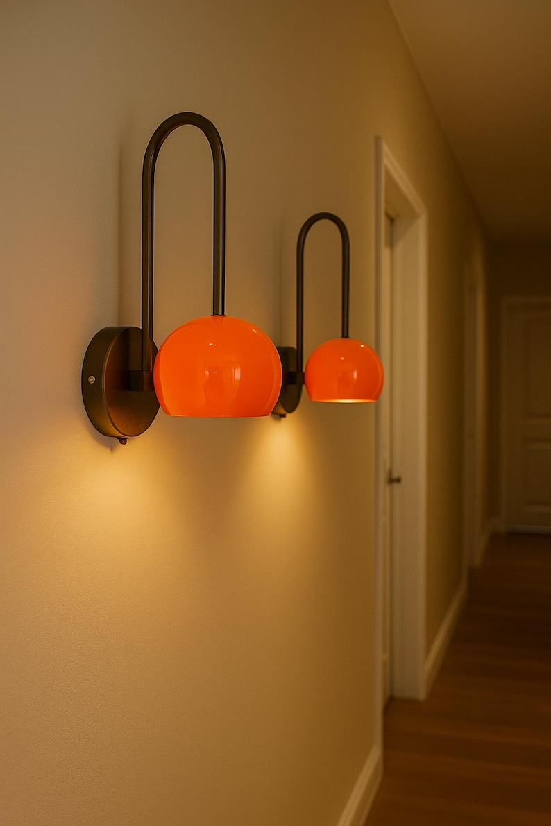 May include: Two modern wall sconces with orange globe shades and dark metal arched arms. The sconces are mounted on a light-colored wall, casting a warm glow. The design is minimalist and stylish, suitable for interior lighting.