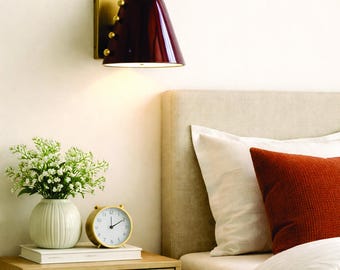 May include: A burgundy wall-mounted lamp with a brass base and accents illuminates a bedroom scene. A nightstand with a vase of white flowers, a gold alarm clock, and a stack of books sits beside a bed with pillows.