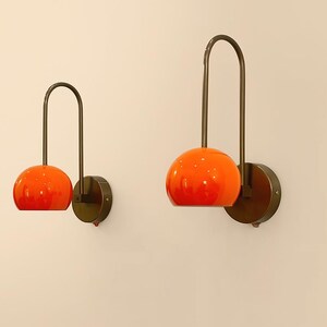 May include: Two orange glass globe wall sconces with dark bronze metal accents. Each sconce features a curved arm and a circular base, creating a modern and stylish lighting fixture. The sconces are mounted on a light beige wall.